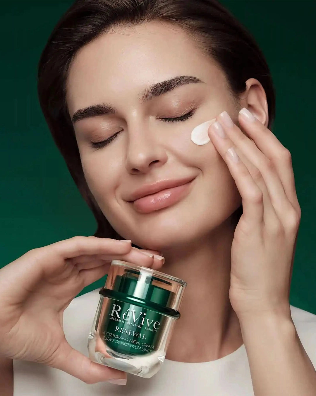 Woman applying cream to her face with a jar of Revive Renewal Cream in front of her.