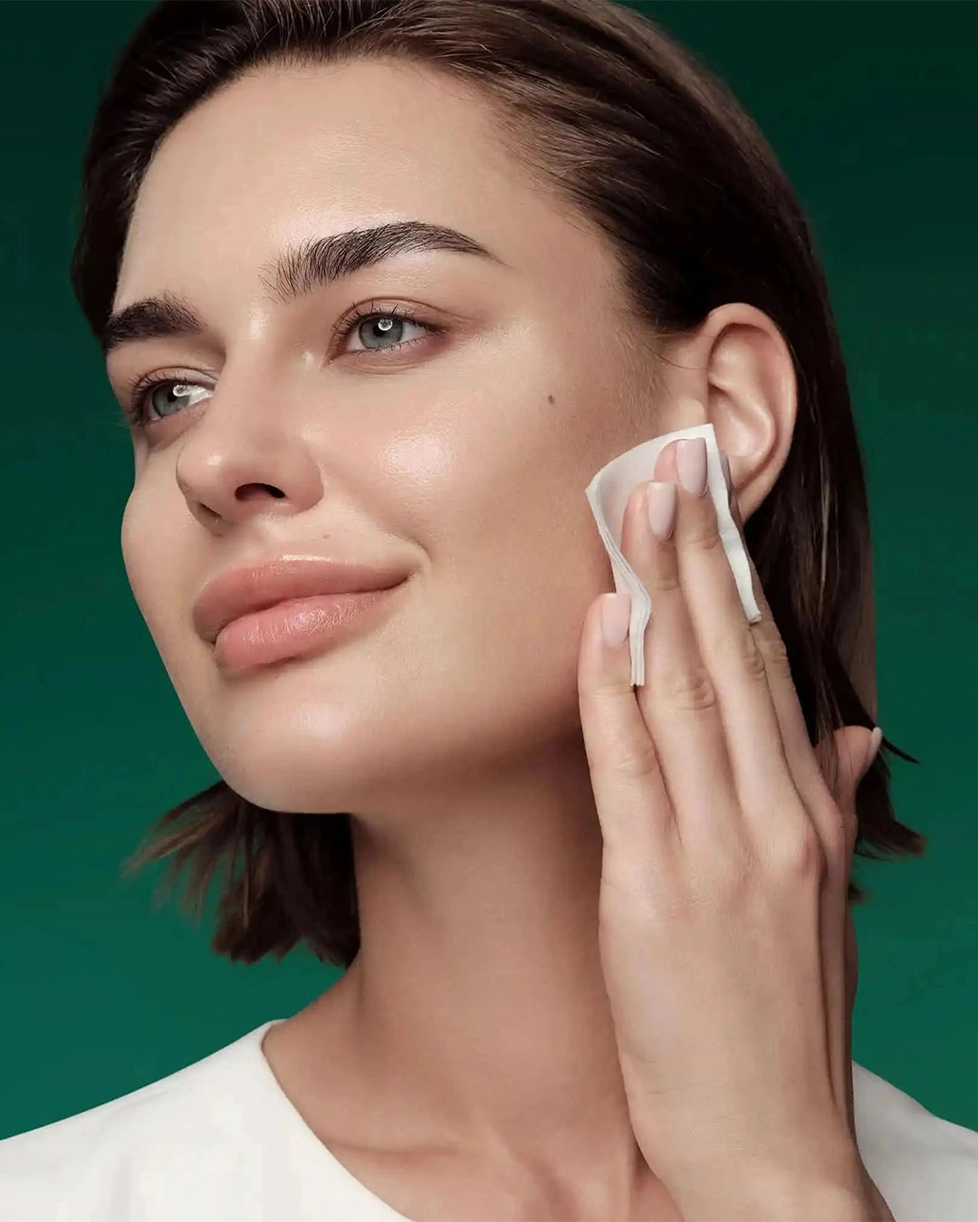 Woman cleaning her face with a cotton pad against a green background