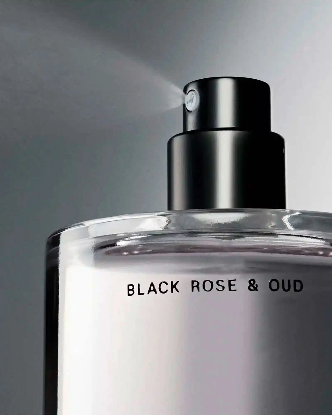 A detailed close-up shot of the top of the Salt & Stone Black Rose & Oud bottle. The image highlights a fine mist being sprayed out from the nozzle of the sleek black atomiser cap, with the product name clearly visible on the glass below.