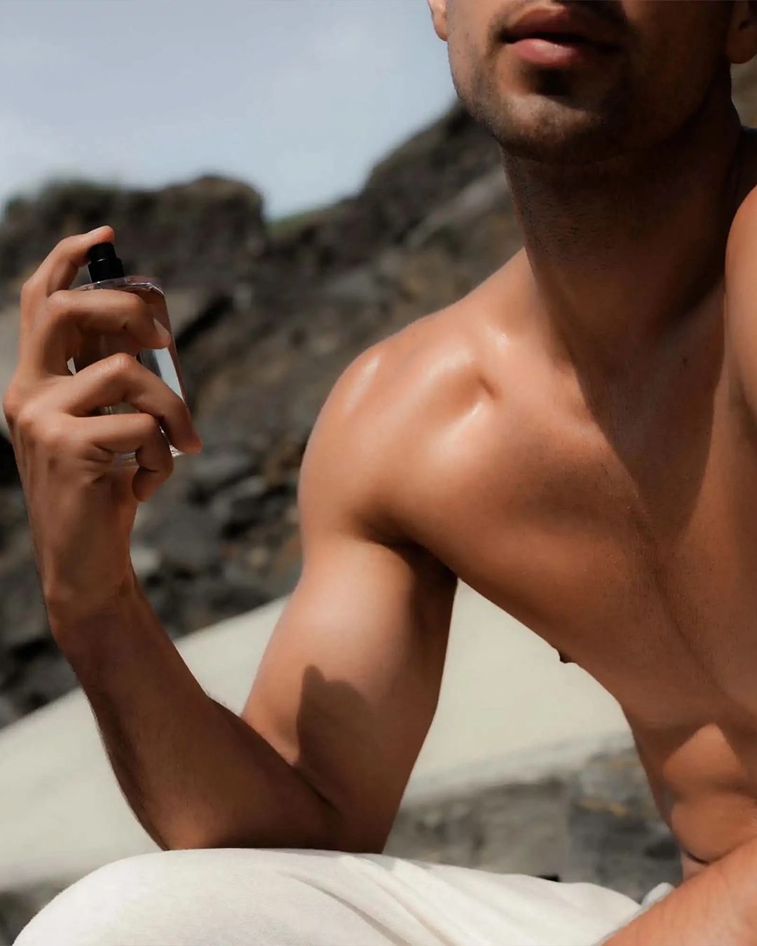 An outdoor lifestyle shot of a muscular, shirtless man holding the Salt & Stone body mist bottle in his hand, preparing to spray it. The blurred background features sunlit coastal rocks.