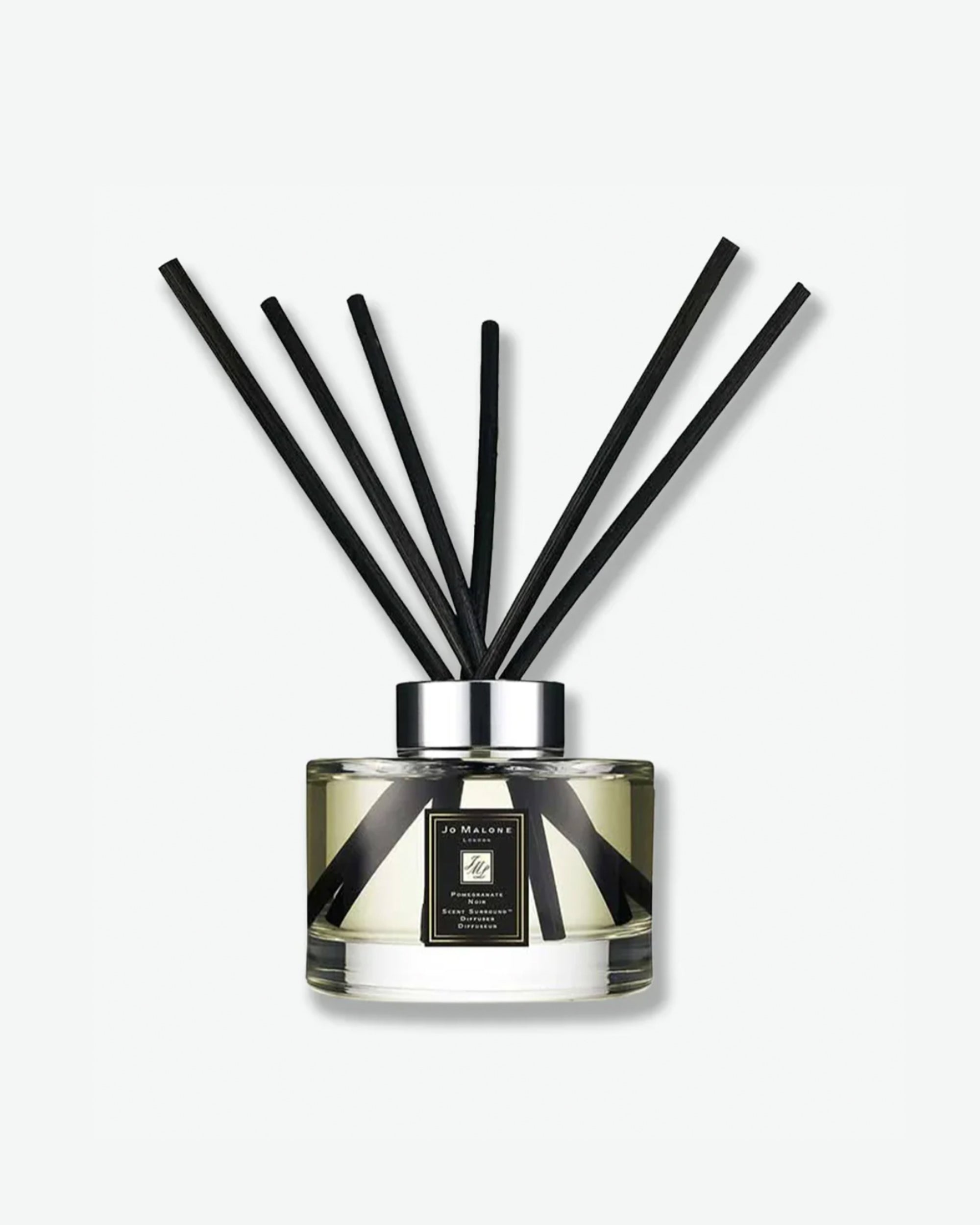 Product shot of a Jo Malone London Pomegranate Noir reed diffuser. The clear, cylindrical glass bottle contains a light amber-tinted fragrance liquid and features the signature square black-and-gold label. Six long, black diffuser reeds fan out symmetrically from the polished silver collar, casting a soft shadow against a clean, minimal white background.
