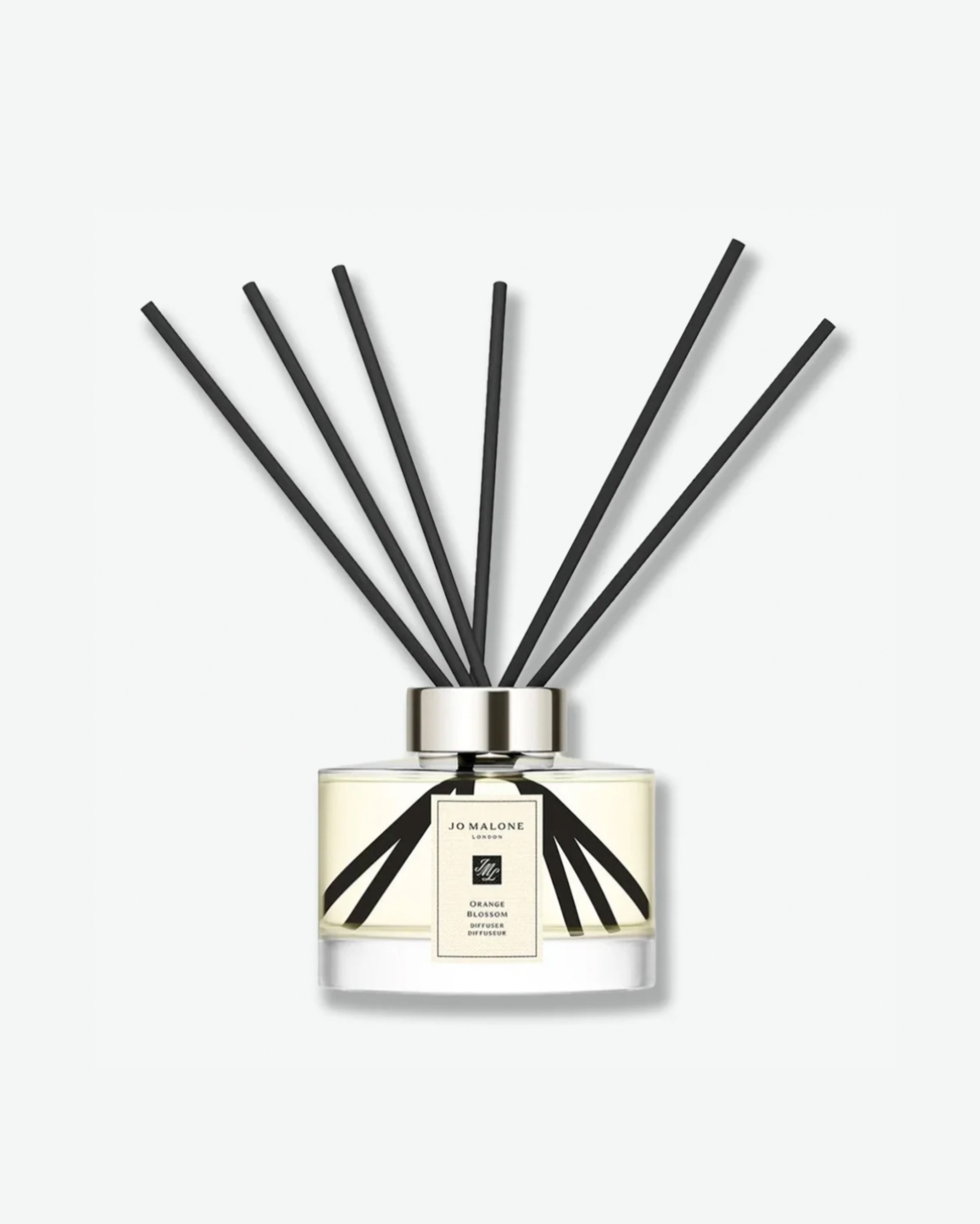Front view of a Jo Malone London Orange Blossom reed diffuser. The clear glass bottle is low, wide, and cylindrical, containing a pale yellow liquid. It is topped with a metallic silver collar from which ten long, slender black diffuser reeds fan out symmetrically upwards. A rectangular cream label on the front features a black border, the brand's name, its monogram logo, and the text "Orange Blossom Diffuser / Diffuseur."