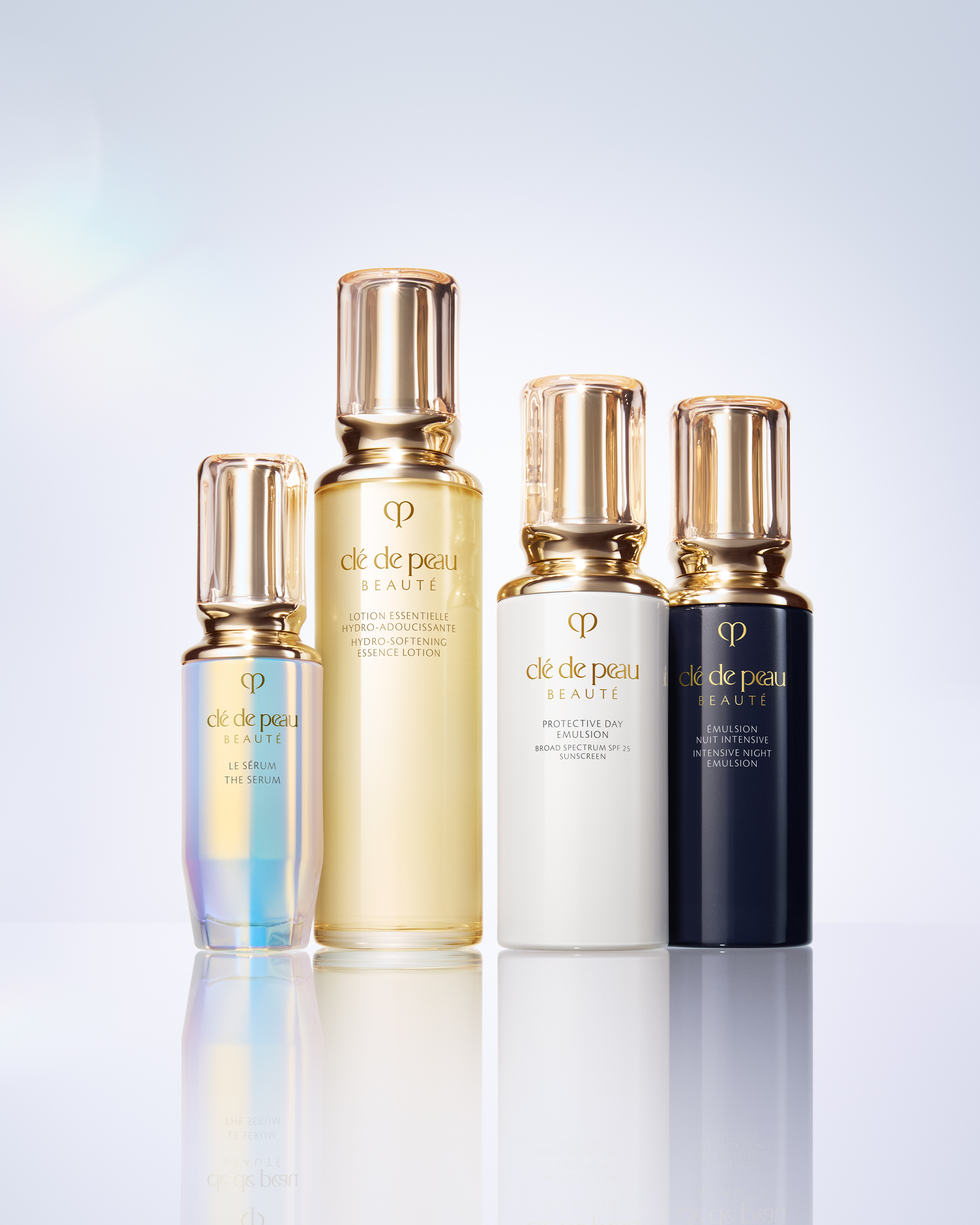 Four skincare bottles from cle de peau BEAUTY arranged in a row on a reflective surface against a light gray gradient background. The bottles are iridescent blue, translucent gold, white, and dark navy, all with shiny metallic gold caps.
