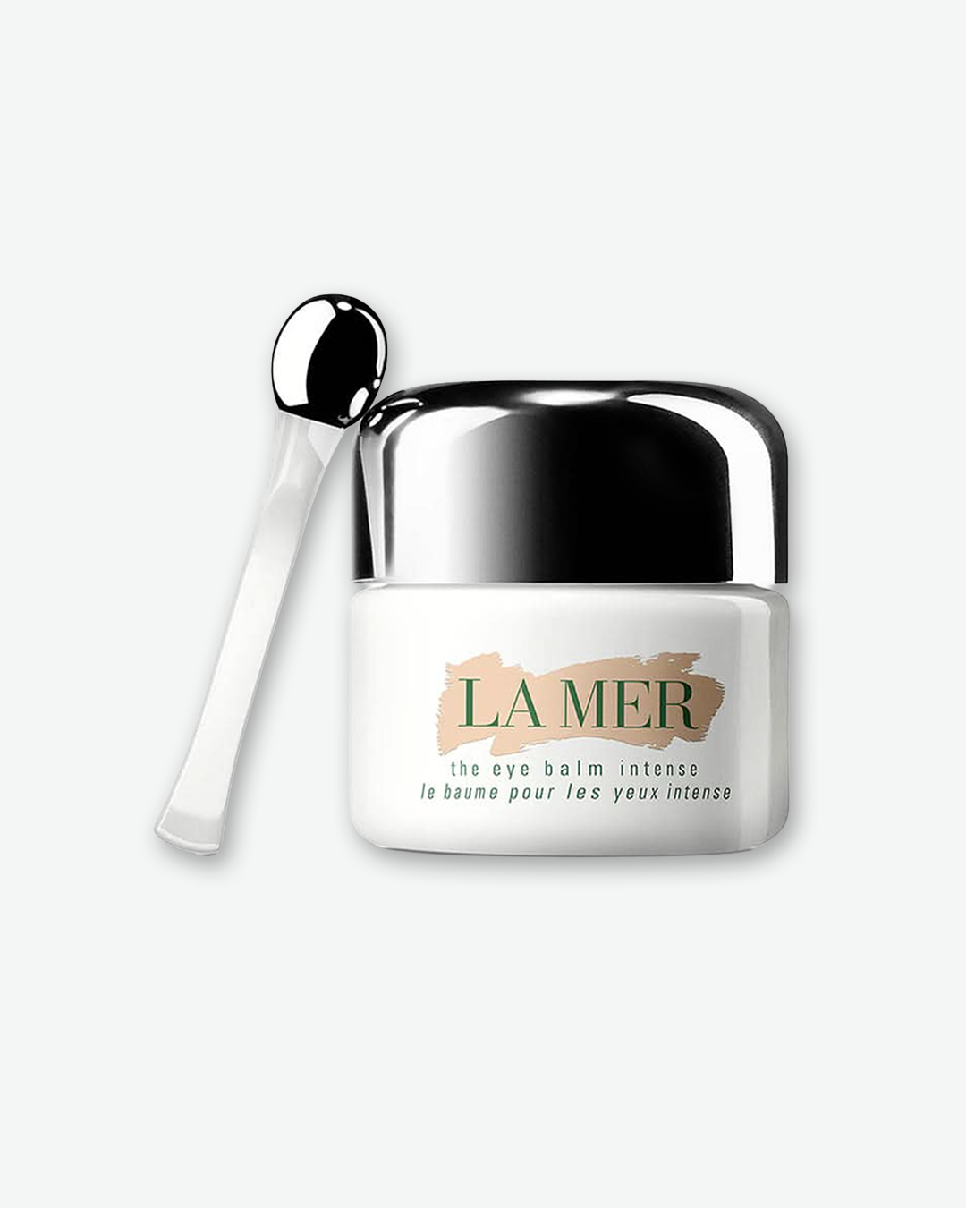 A product shot of a La Mer The Eye Balm Intense jar next to its applicator tool, centered against a plain light gray background. The small, solid white glass jar features a highly reflective chrome silver dome cap and a gold-brushed logo section on the front. Leaning vertically against its left side is a clear acrylic spatula applicator tool with a polished silver ball tip.