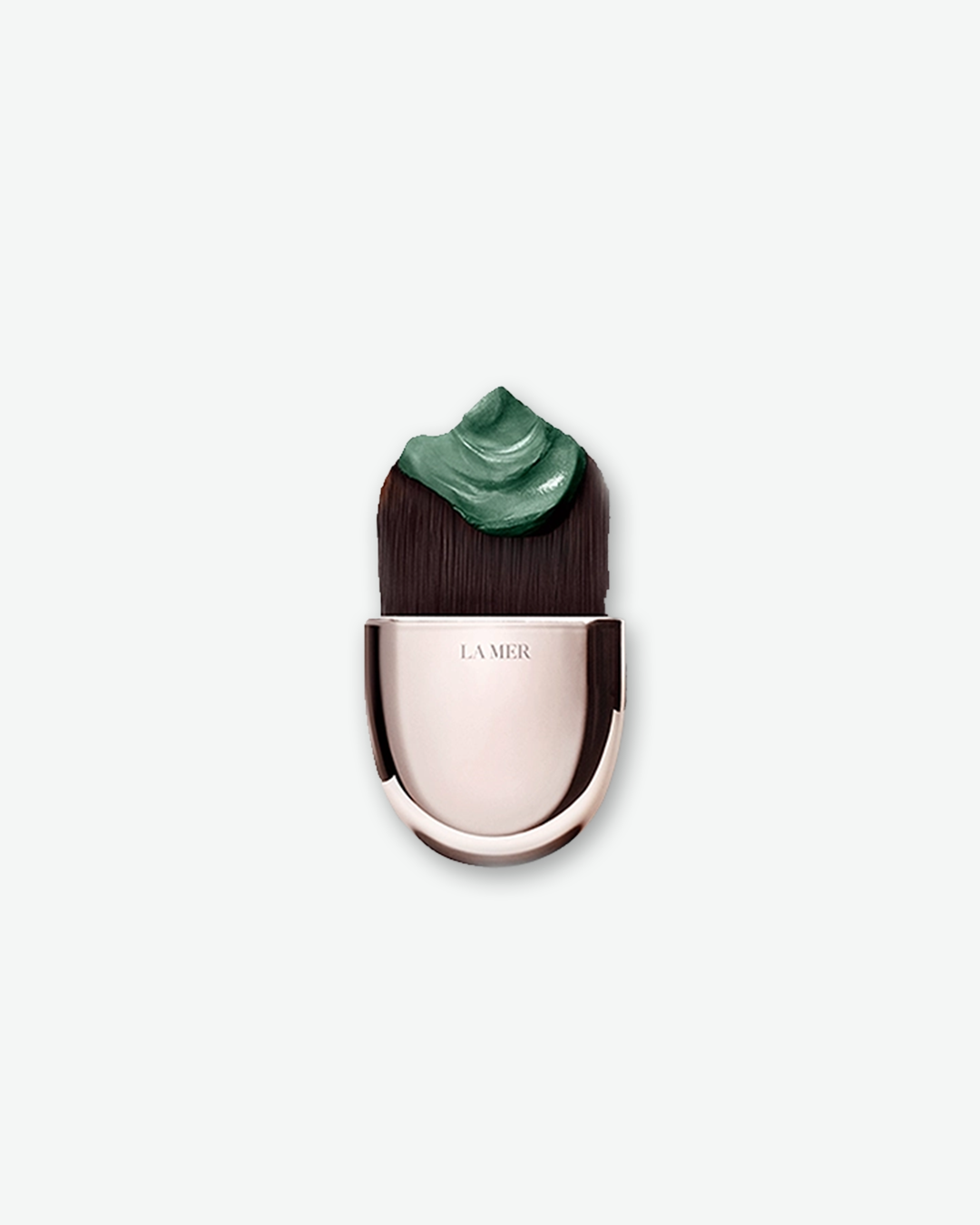 A product shot of a La Mer facial brush, centered against a plain light gray background. The applicator features a smooth, metallic champagne-gold handle. Its dense, dark brown bristles are cut into a wide, flat profile, holding a thick dollop of a dark emerald-green cream mask on top.