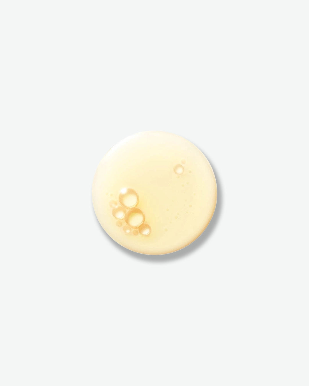 A top-down view of a single round droplet of golden-yellow skincare liquid. The texture shows slight transparency with a cluster of tiny bubbles on one side, casting a soft shadow on a clean, minimal light gray background.