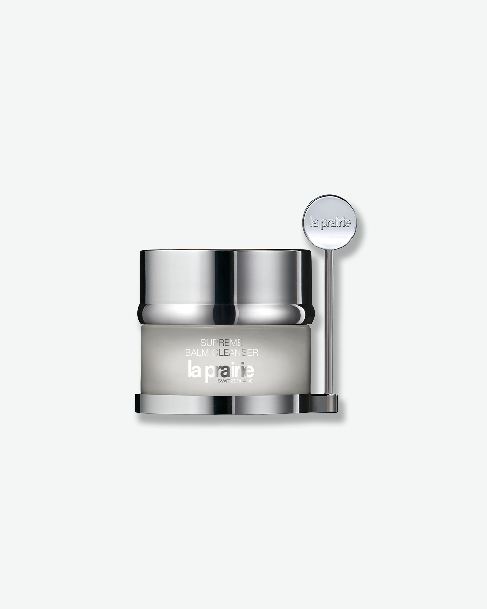 Product shot featuring a La Prairie Supreme Balm Cleanser jar with its companion spatula against a plain gray background. The frosted clear glass jar is mounted onto a polished silver metallic base that holds a vertical, matching silver spatula with a round handle head embossed with the brand name.