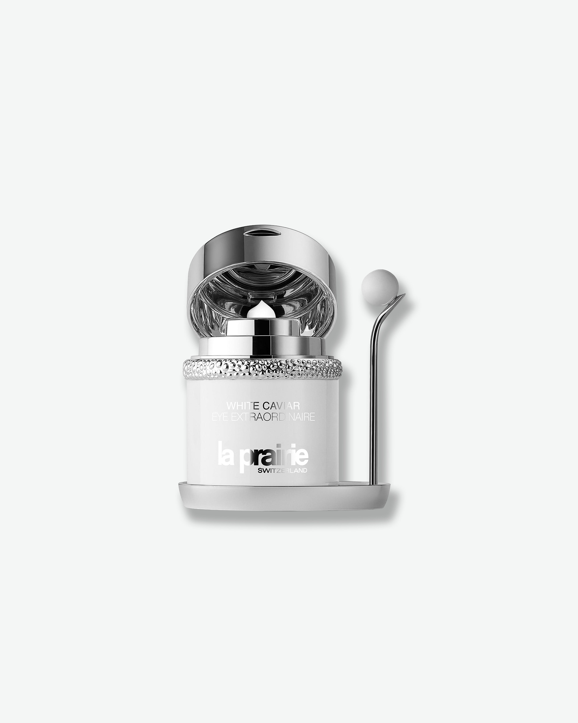 Product shot of the La Prairie White Caviar Eye Extraordinaire jar shown with its silver lid flipped open. The interior features a reflective silver pump mechanism with a central dispensing aperture, while the white ceramic massage wand remains docked in its holder to the right.