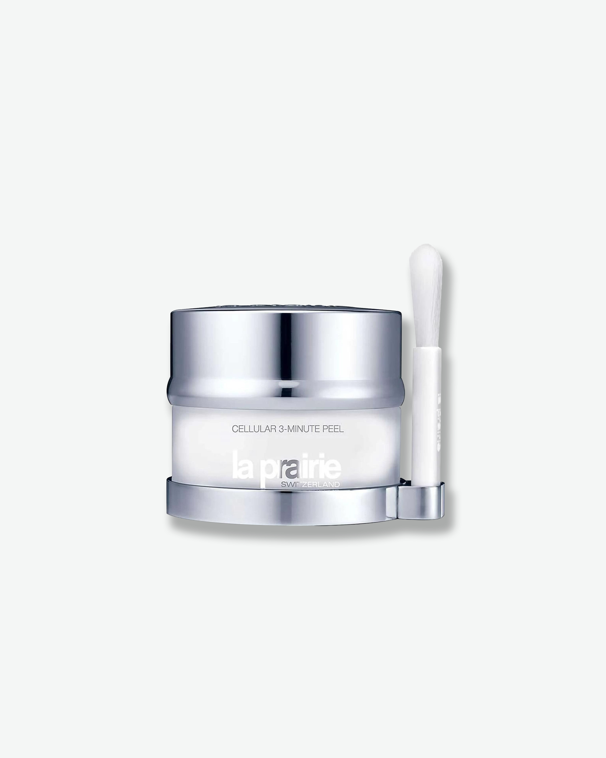 Product shot featuring a La Prairie Cellular 3-Minute Peel jar with its companion application brush. The frosted white glass jar is nestled on a polished silver base that includes a matching vertical holder on the right side for its sleek white applicator brush.