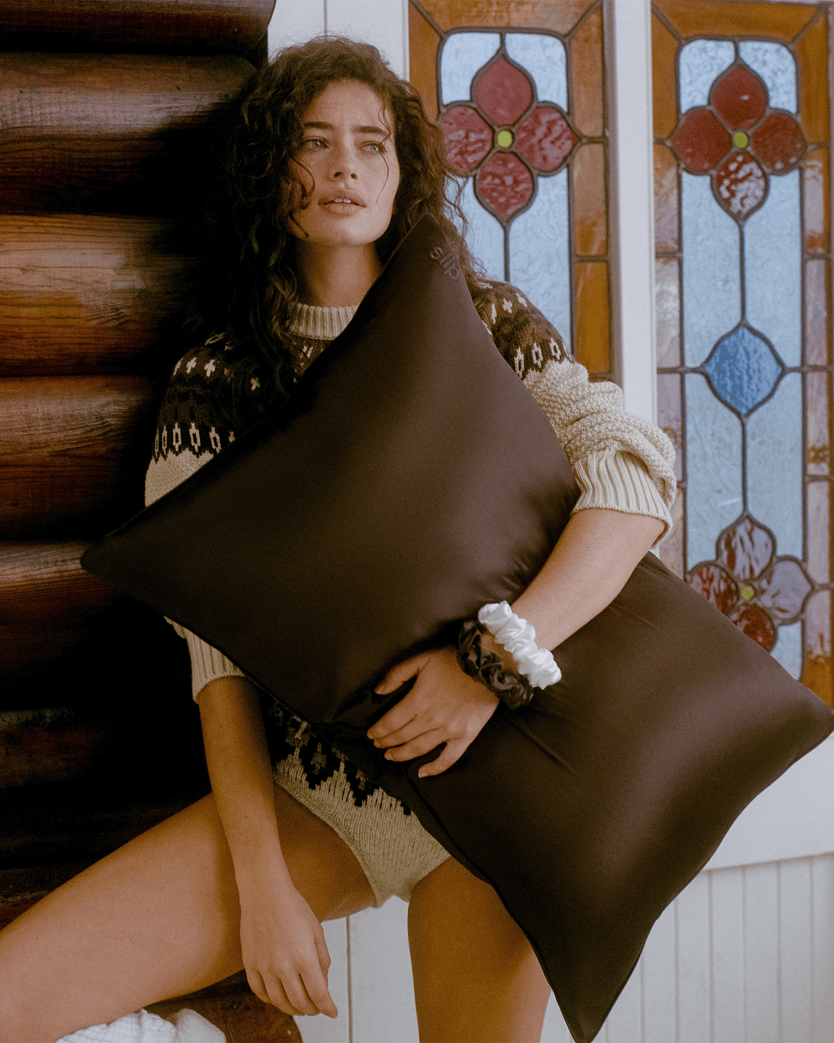 Woman with curly dark hair wearing a cozy patterned sweater and holding an oversized chocolate brown Slip silk pillowcase in a rustic cabin setting.
