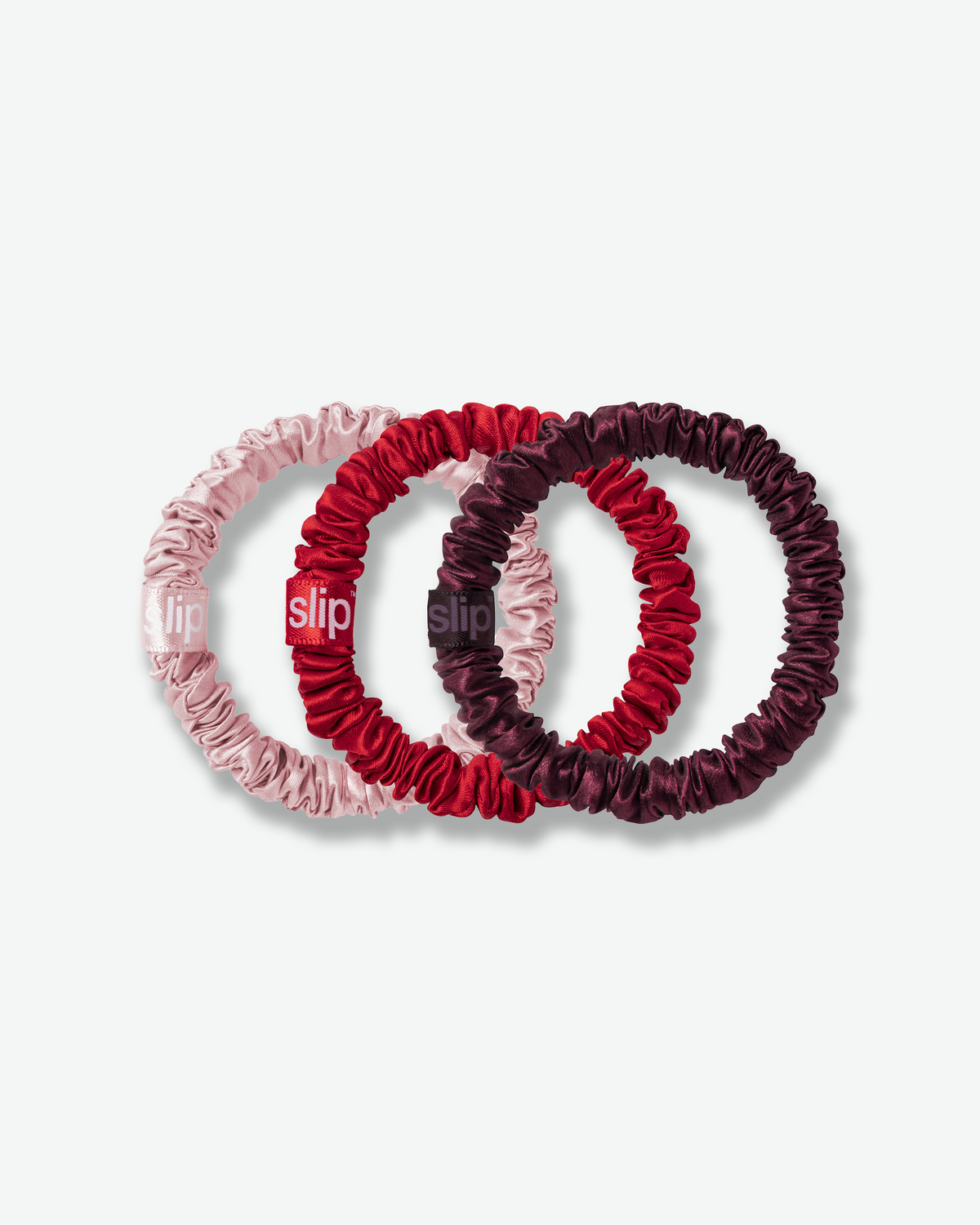 Slip silk skinny scrunchies in blush pink, bright red, and burgundy, featuring smooth silk fabric and branded tags on a light gray background.