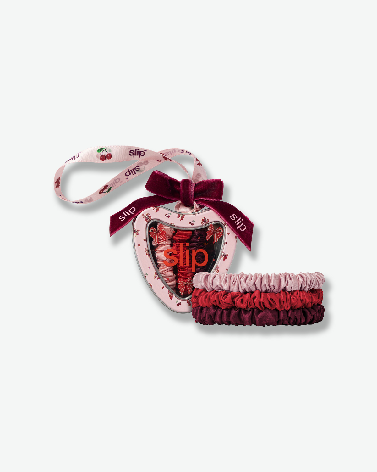 Slip holiday silk skinny scrunchies in pink, red, and burgundy, displayed alongside a heart-shaped gift ornament decorated with cherries, bows, and a burgundy velvet ribbon.