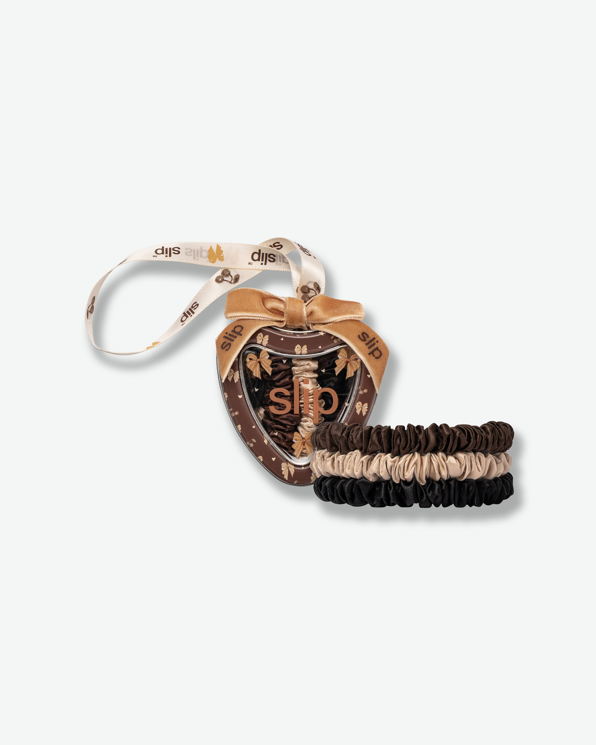 Slip silk skinny scrunchies in chocolate brown, champagne, and black, displayed beside a festive heart-shaped ornament with a gold bow and Slip-branded ribbon.