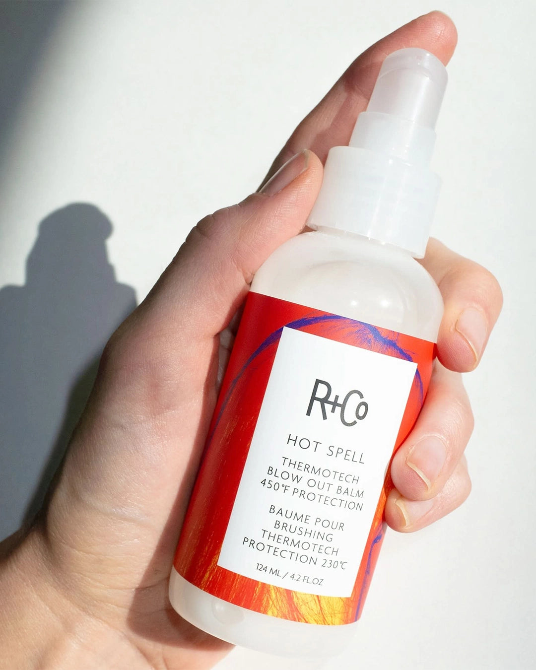 Hand holding a bottle of R+Co Hot Spell Thermotech product against a white background