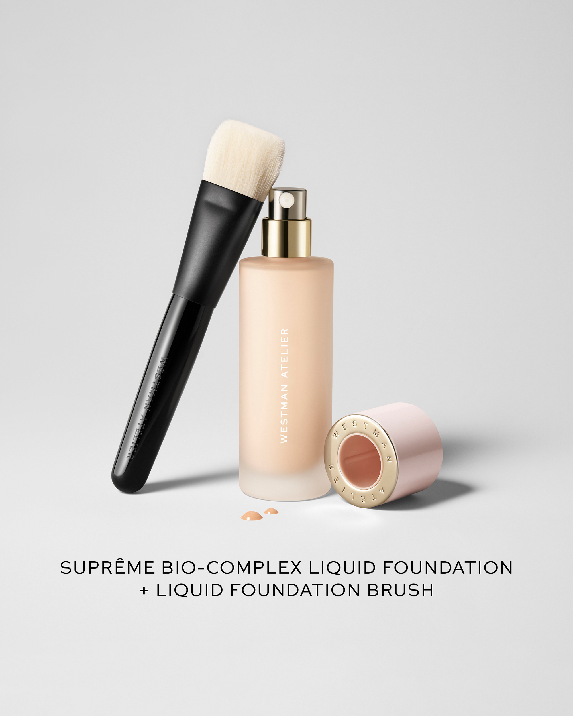 Westman Atelier Suprême Bio-Complex Liquid Foundation bottle shown next to its matching liquid foundation brush with light bristles and a pink cap lying nearby.