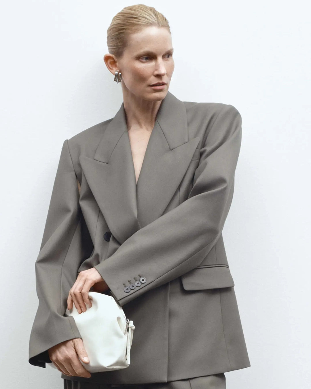 Woman wearing a gray suit holding a white clutch against a plain background