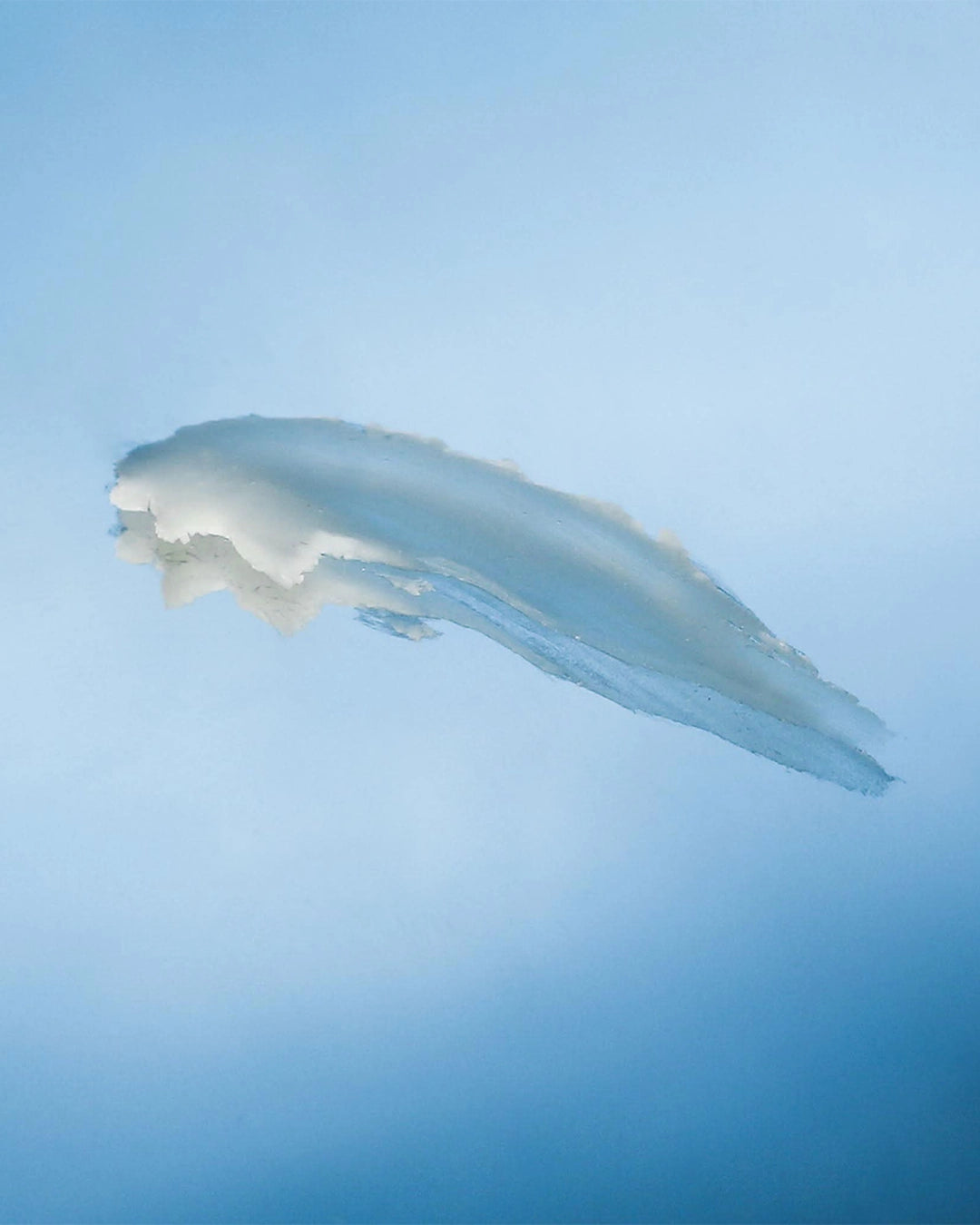 A swatch of translucent balm is displayed against a soft blue gradient background. The semi-sheer formula is spread in a smooth, sweeping stroke, revealing its rich, cushiony texture and glossy finish. The image highlights the product’s nourishing consistency and lightweight, moisturizing appearance.
