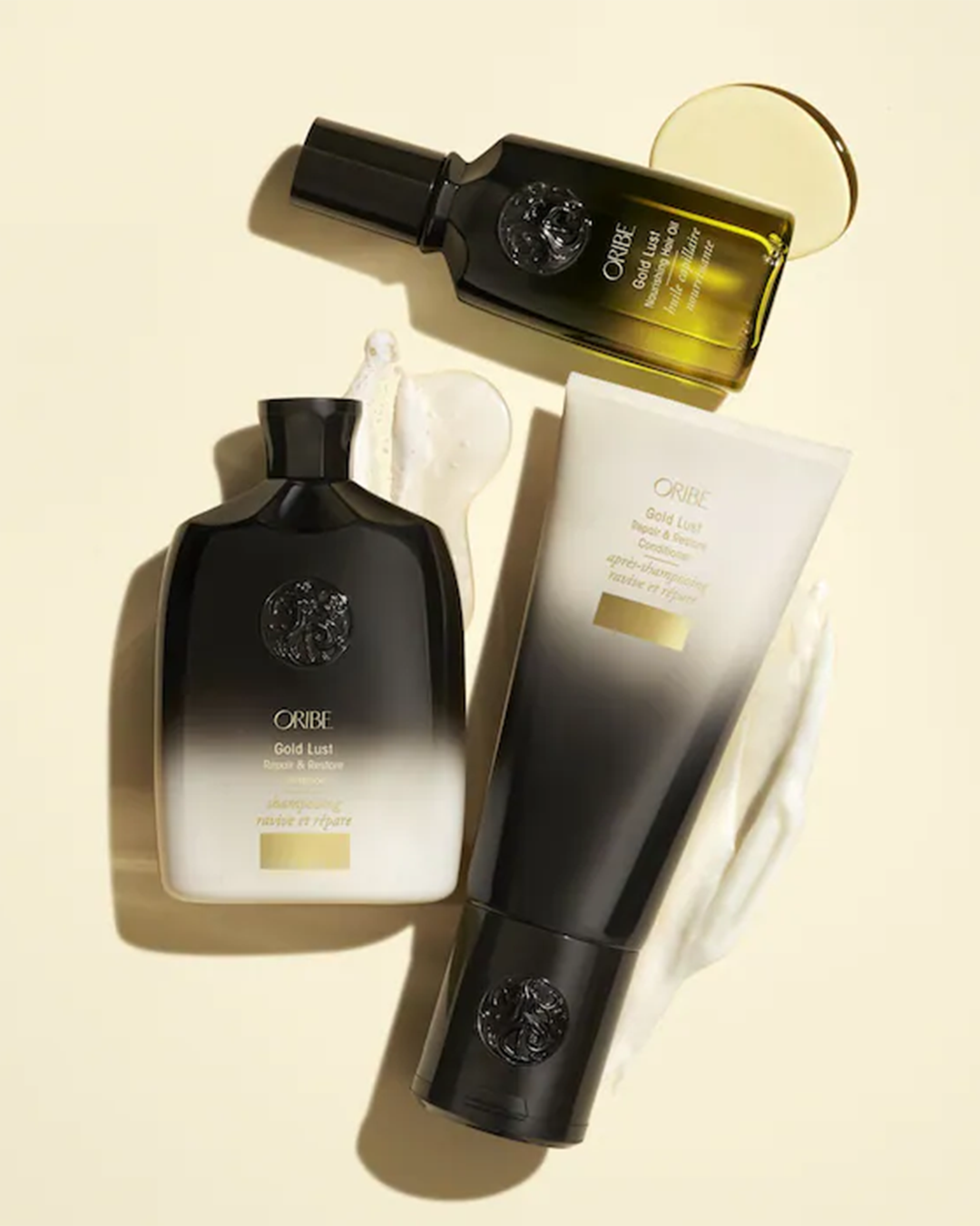 An overhead flat lay of Oribe haircare products on a warm, pale yellow background. It includes a bottle of Gold Lust Shampoo, a tube of Gold Lust Conditioner with a stroke of thick white cream beside it, and a bottle of Gold Lust Nourishing Hair Oil with a drop of golden oil pooled nearby.