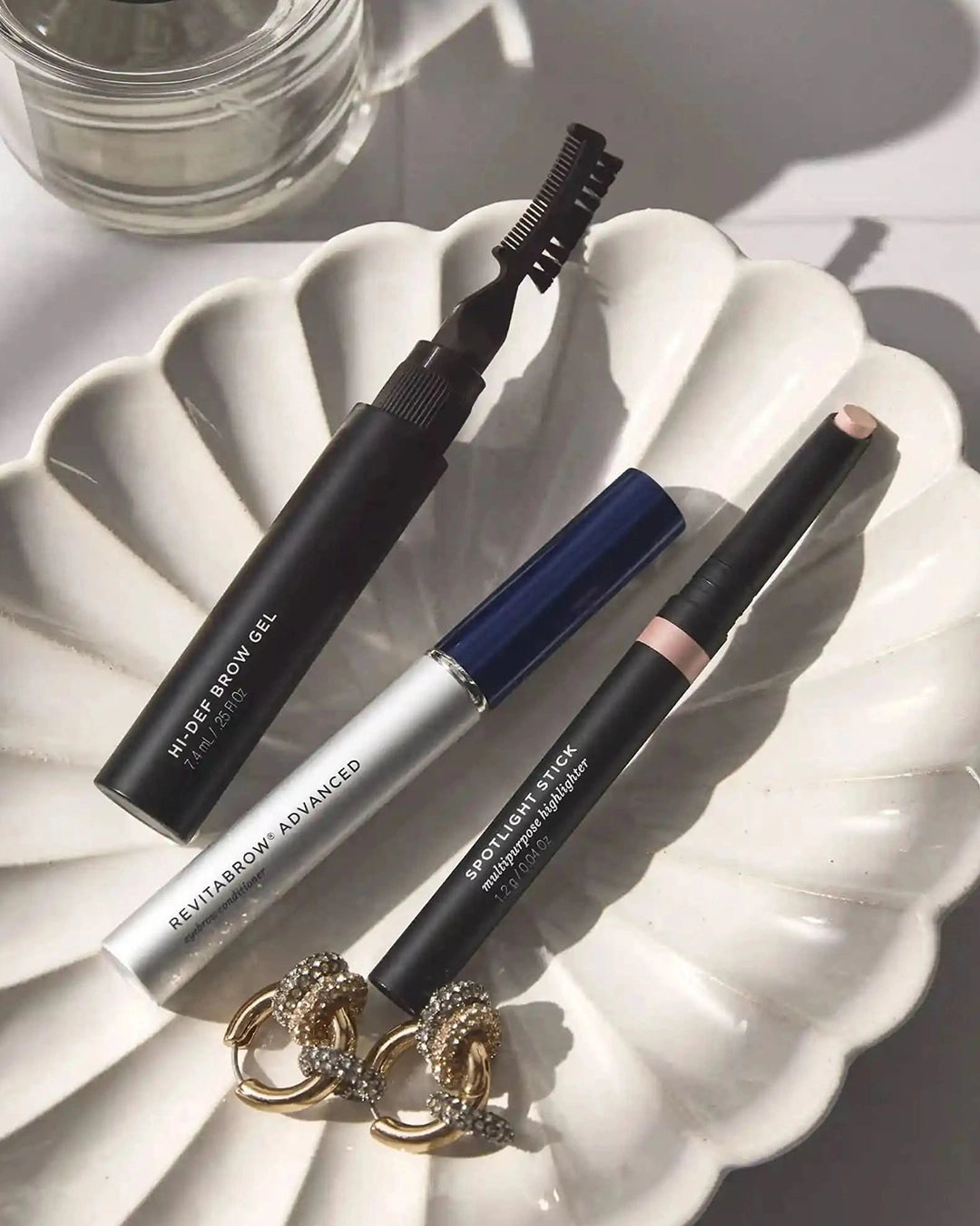 Three makeup products on a white decorative plate with a glass container in the background.