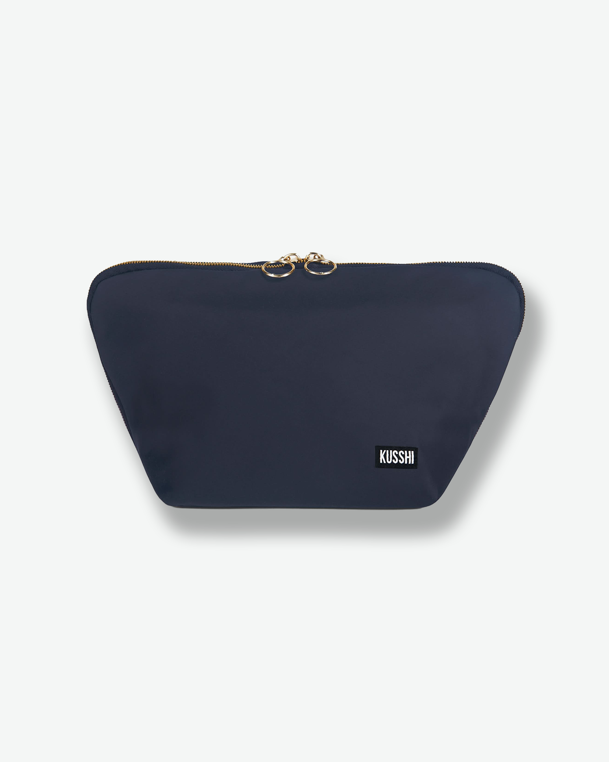 Front view of the large navy blue fabric KUSSHI makeup bag shown completely zipped shut. The top zipper track meets at the center with two polished gold circular ring zipper pulls.
