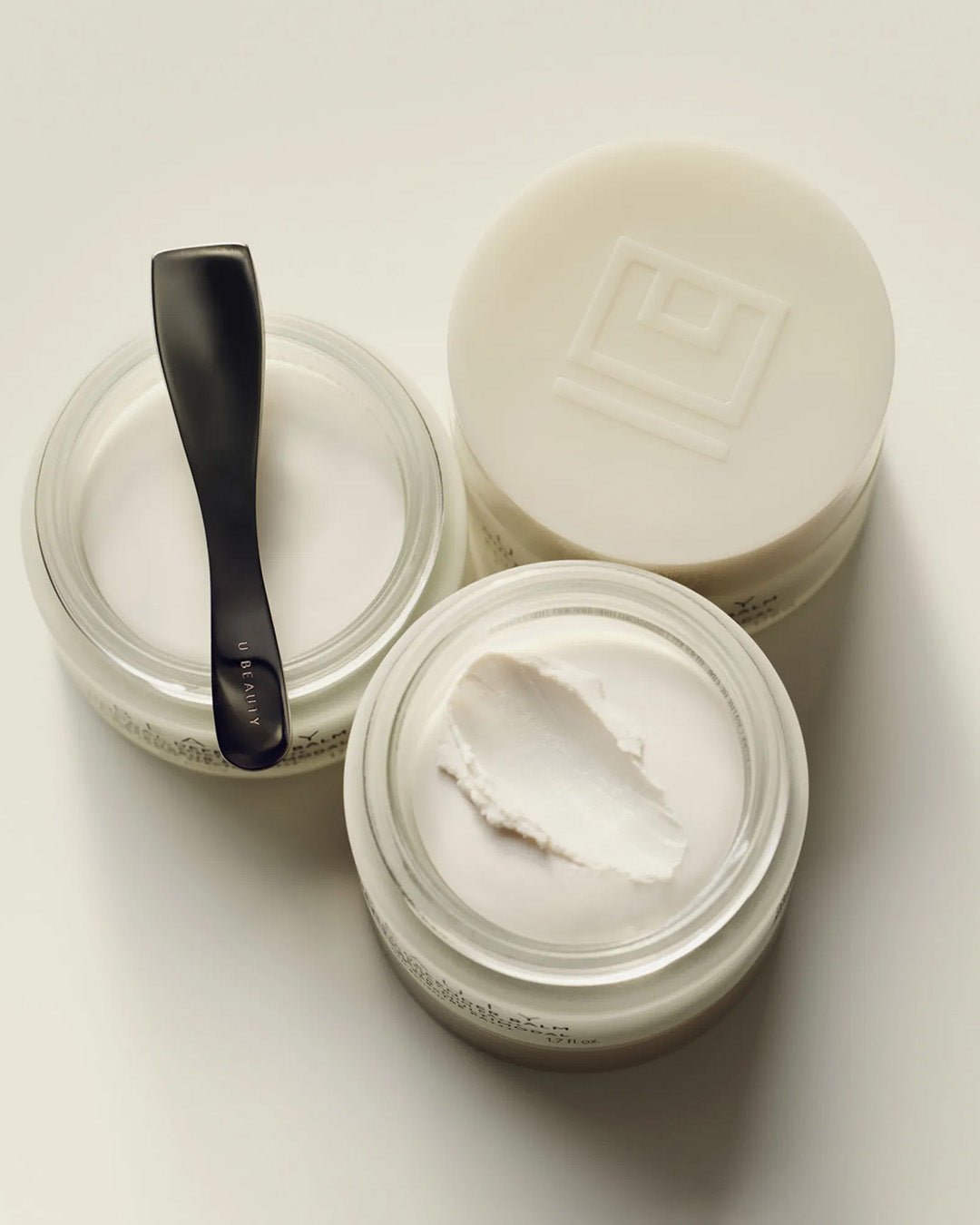 Two jars of cream with a black spoon on a light background