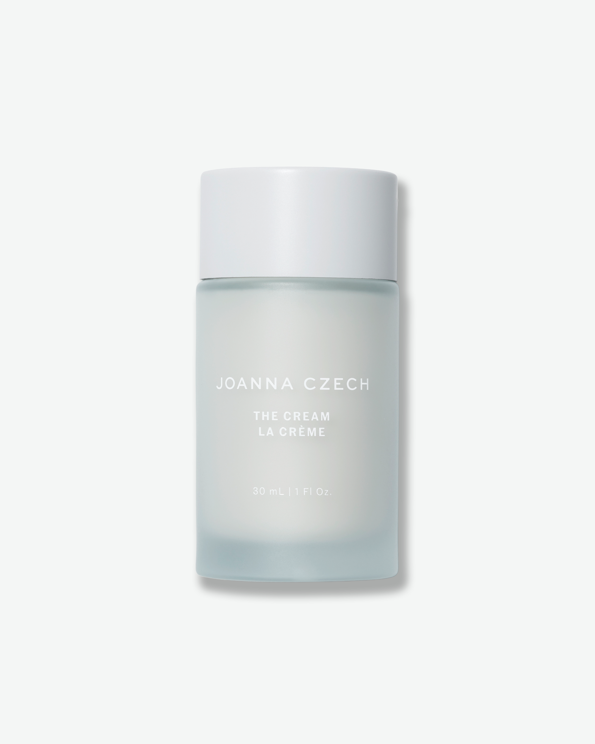 Front view of a Joanna Czech "The Cream / La Crème" bottle centered against a plain gray background. The minimalist container features a frosted, light ice-blue glass body printed with subtle white text, topped with a smooth, matte white cylindrical cap.