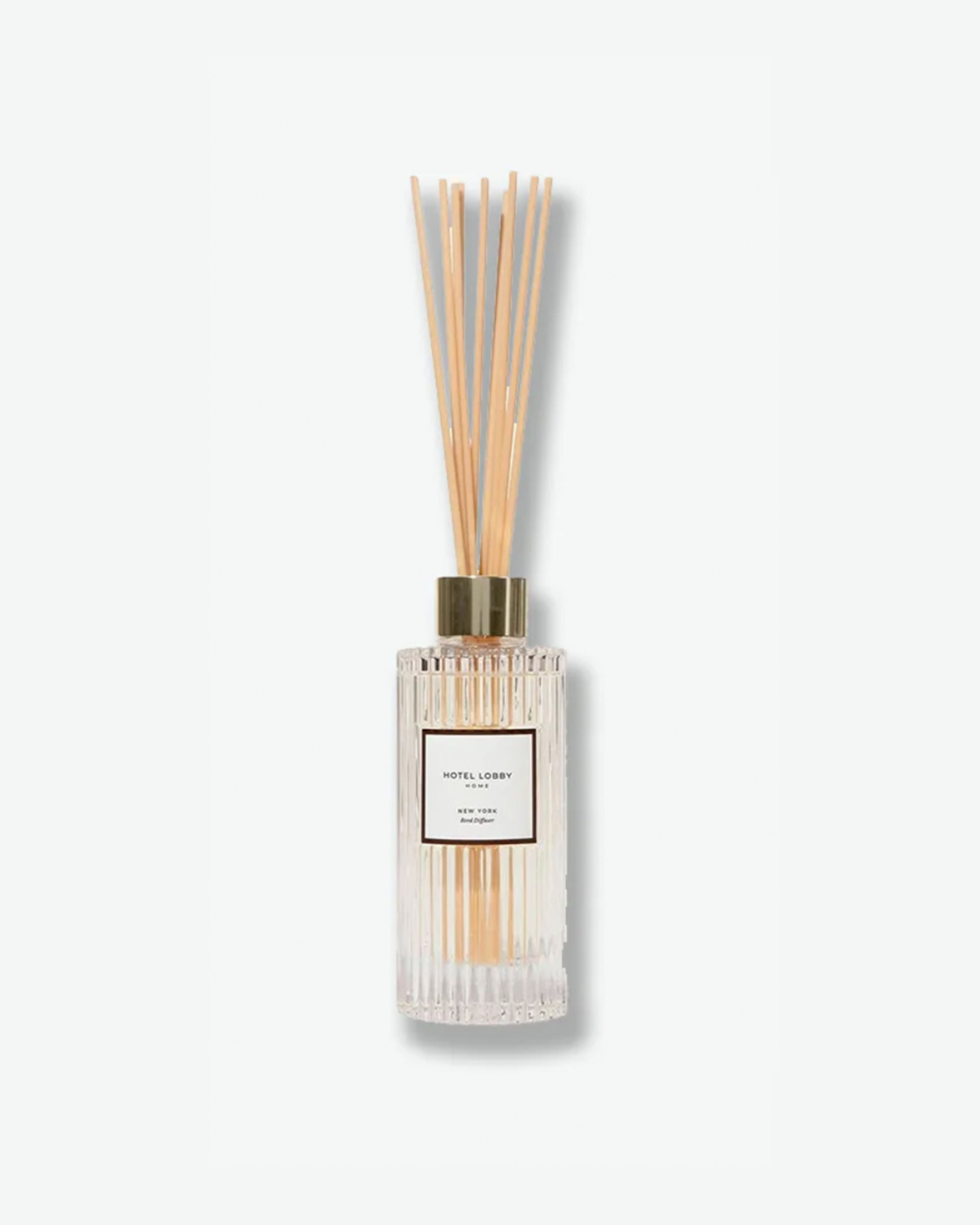 Hotel Lobby Home New York Reed Diffuser featuring a fluted clear glass bottle filled with clear liquid and light wooden reeds extending upward, isolated against a light grey background.