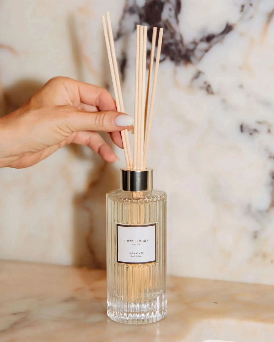 Hand holding a diffuser stick over a clear bottle with a label against a marble background