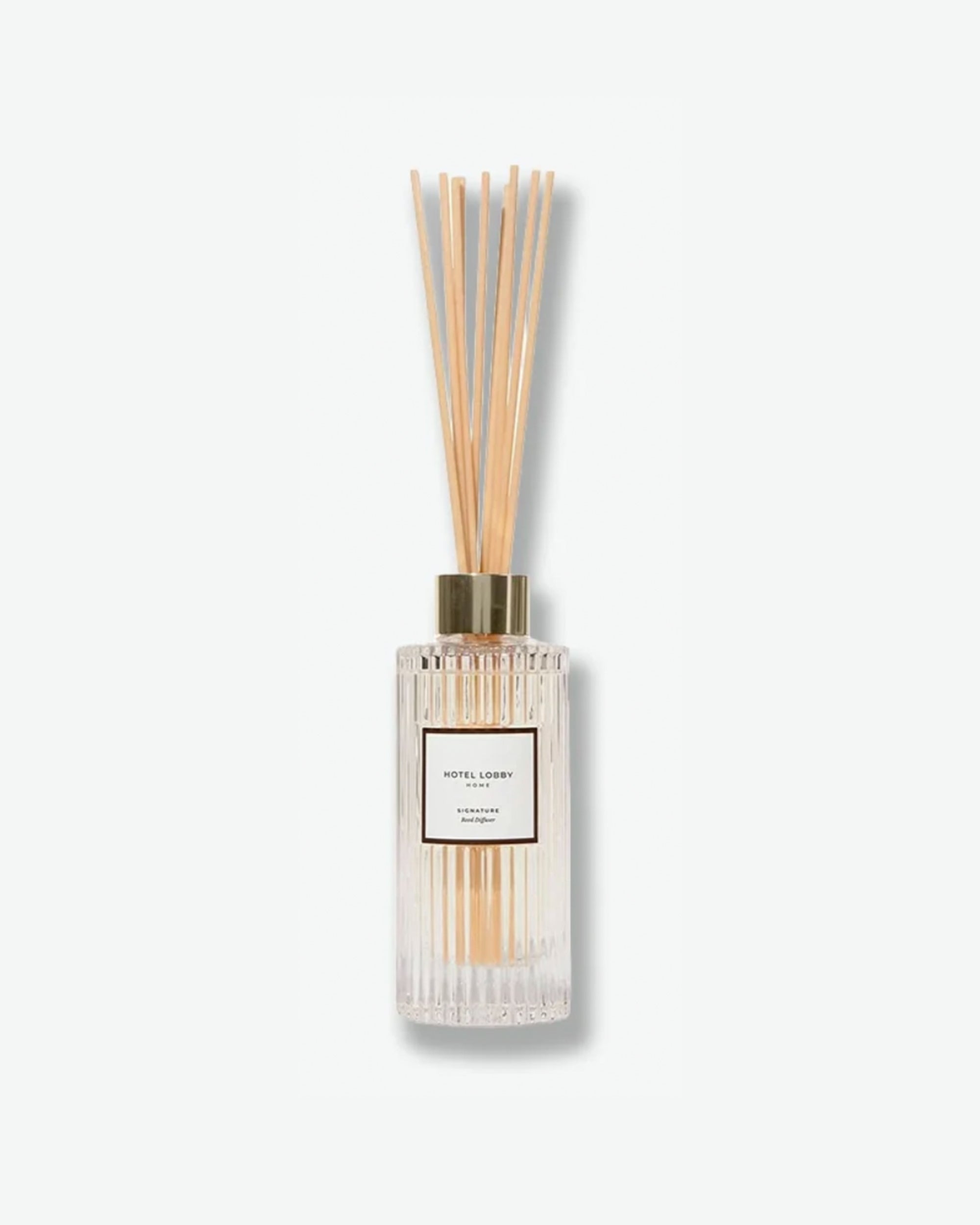 Hotel Lobby Home Signature Reed Diffuser featuring a fluted clear glass bottle filled with clear liquid and light wooden reeds extending upward, isolated against a light grey background.