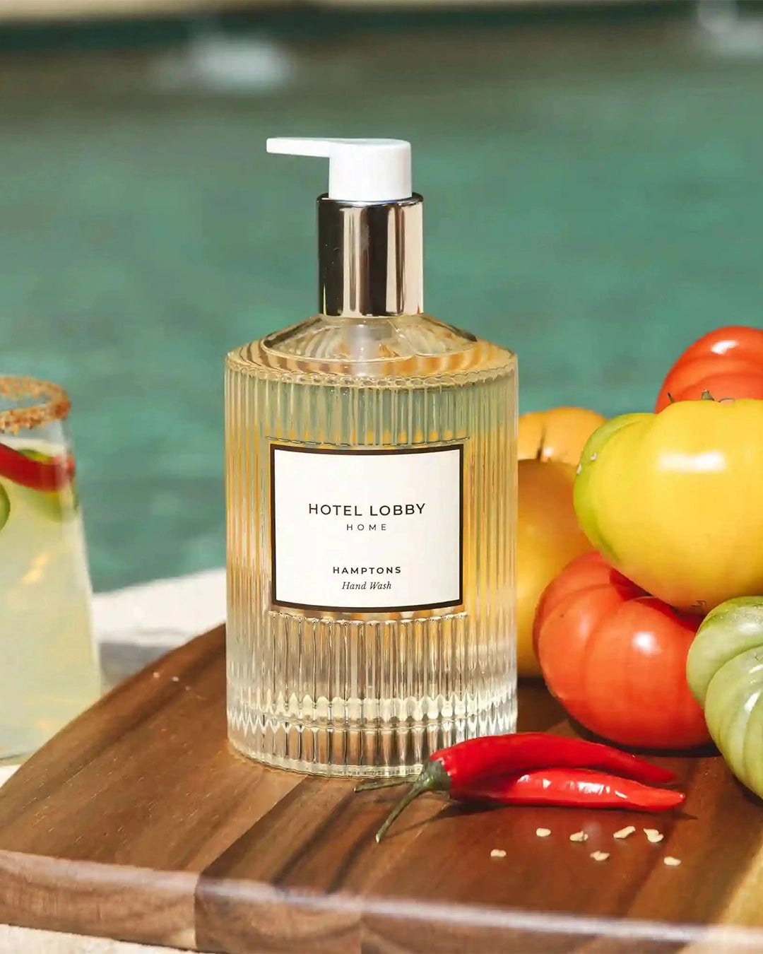 Glass bottle with pump labeled 'Hotel Lobby' on a wooden surface with fruits and a drink.