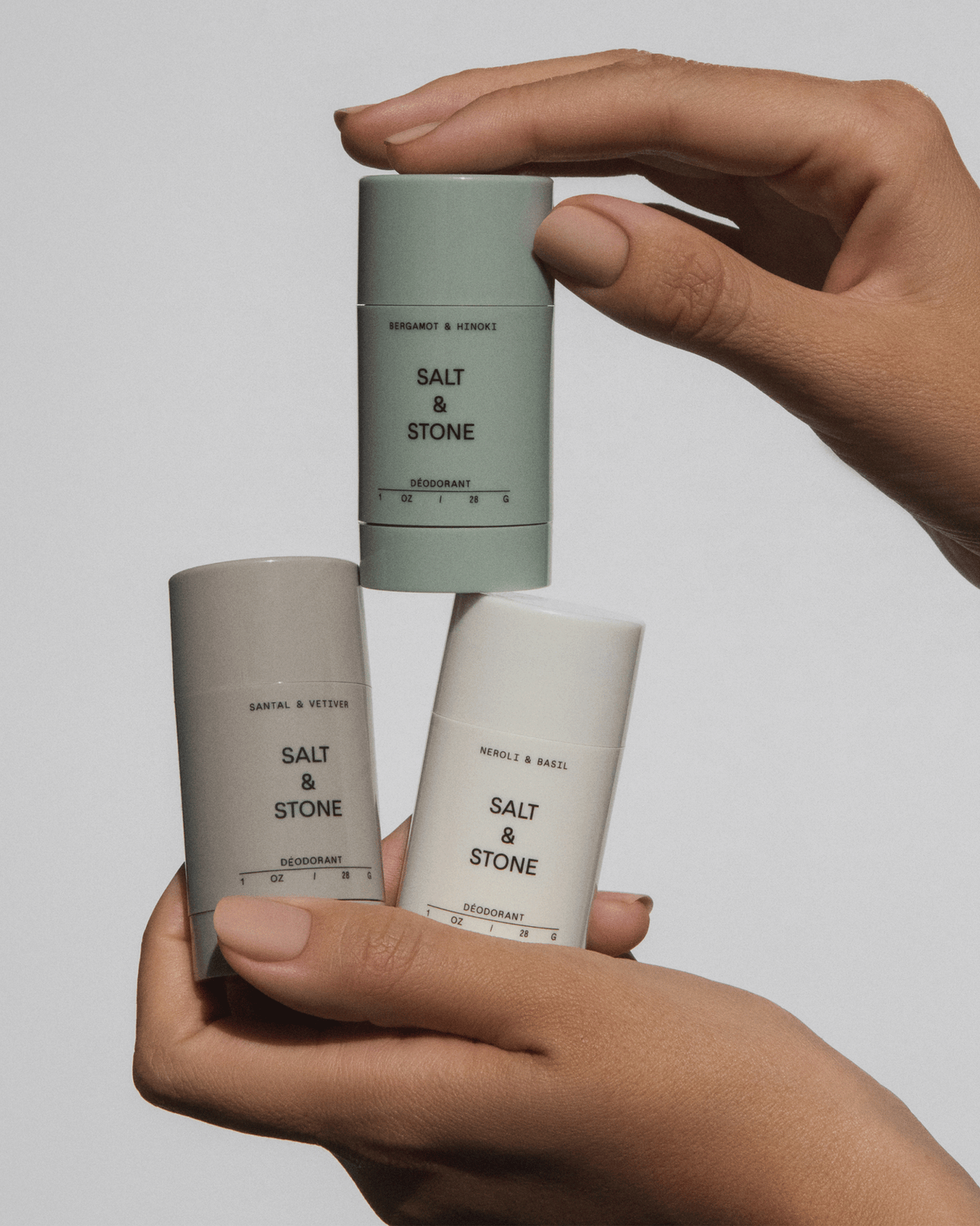 Hands balancing three small Salt & Stone deodorant sticks in grey, sage green, and cream against a light grey background.