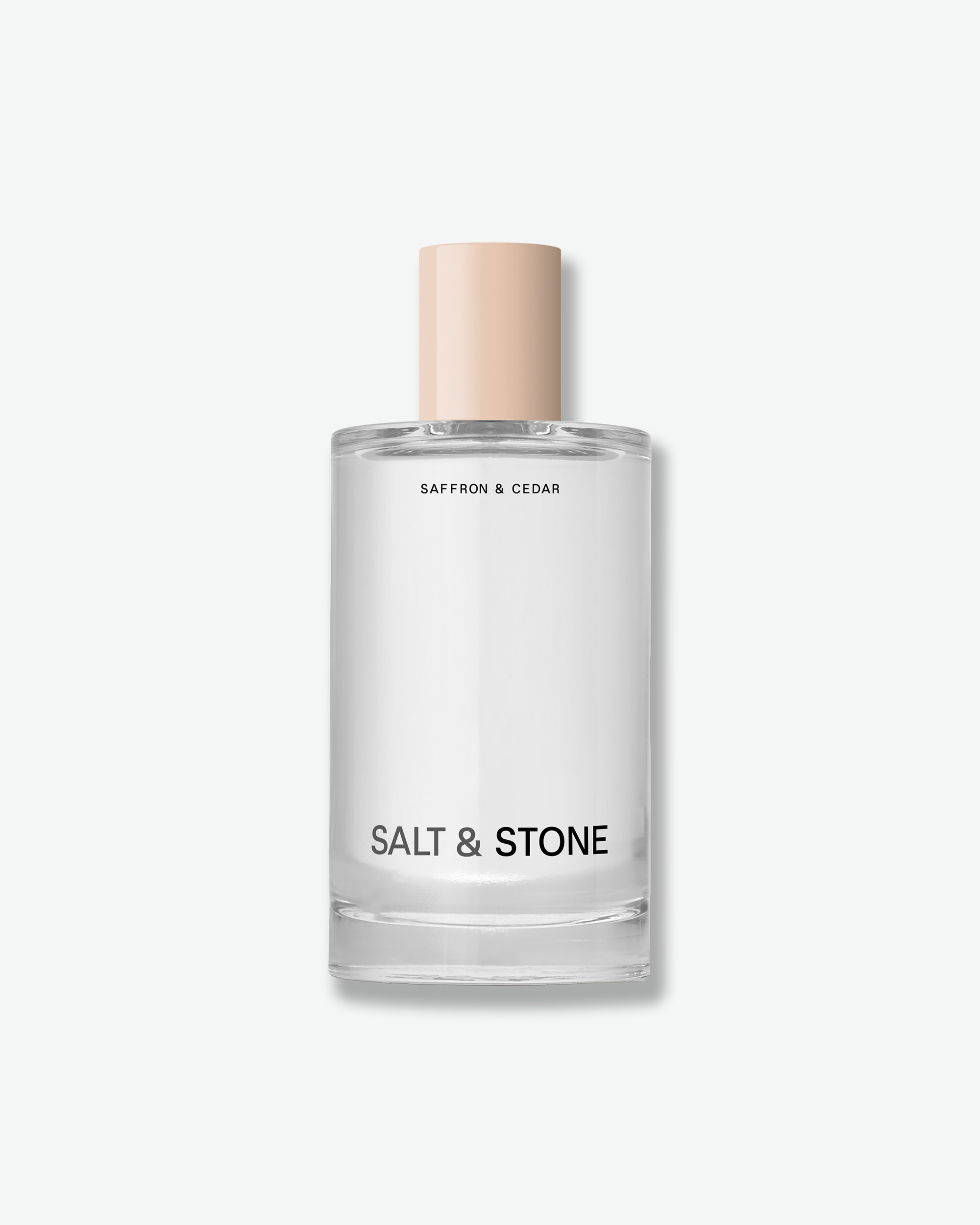 A clear glass perfume bottle with a pale peach cap, labeled 'SAFFRON & CEDAR' and 'SALT & STONE', centered on a light gray background.