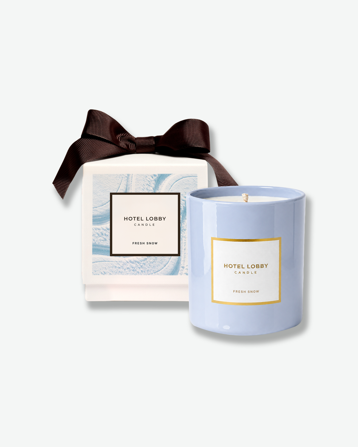 Hotel Lobby Candle Fresh Snow in a light blue ceramic vessel, displayed alongside its white gift box featuring icy blue artwork and a dark brown ribbon bow.