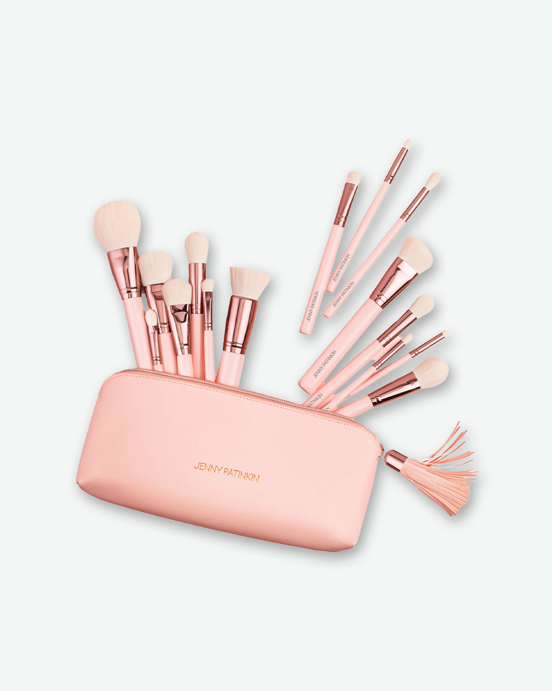 A product shot of a collection of makeup brushes emerging from a pink cosmetic case against a plain light gray background. The brushes feature light pink handles, shiny rose-gold ferrules, and soft white bristles. The pink saffiano leather pouch features the name "JENNY PATINKIN" in small gold lettering on the front and has a matching pink fringe tassel attached to the rose-gold zipper.