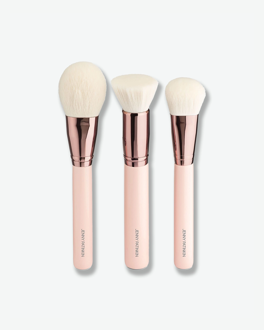 A product shot displaying three large complexion makeup brushes standing upright against a plain light gray background. The brushes have thick, light pink handles with gold brand lettering, metallic rose-gold ferrules, and dense white bristles—one powder brush, one flat-top buffing brush, and one tapered blush brush.