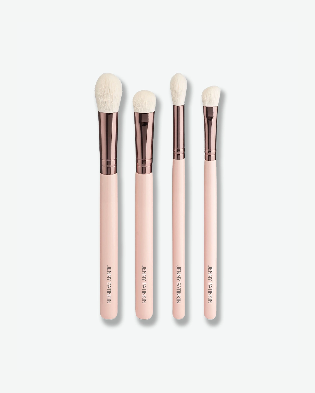 A product shot displaying four makeup brushes standing vertically in a row against a plain light gray background. The brushes feature light pink handles with "JENNY PATINKIN" printed vertically near the base, glossy rose-gold ferrules, and soft white bristles of varying shapes and densities for the face and eyes.