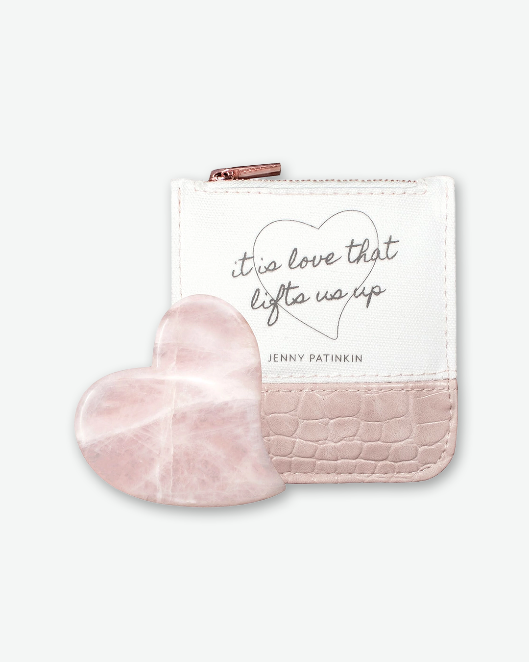 A product shot of a rose quartz gua sha tool leaning against a square storage pouch, centered on a light gray background. The gua sha stone is carved into a smooth, polished heart shape with a soft pink color and subtle natural white veining. The pouch behind it is cream-colored cotton fabric on the top half with the script text "it is love that lifts us up" above "JENNY PATINKIN", and a pink crocodile-textured faux leather on the bottom half, finished with a rose-gold top zipper.