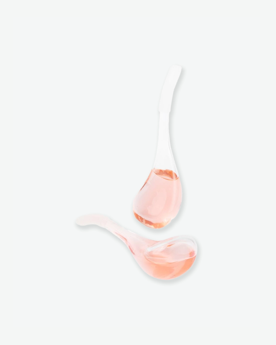 A product shot displaying two glass facial cryo globes filled with a light pink liquid, artistically arranged on a plain light gray background. The globes are floating at an angle; one stands semi-upright showing its tear-shaped bulb filled with fluid and its frosted handle extending upward, while the second globe lies horizontally below it, showing the curved contours of its glass cooling bulb.