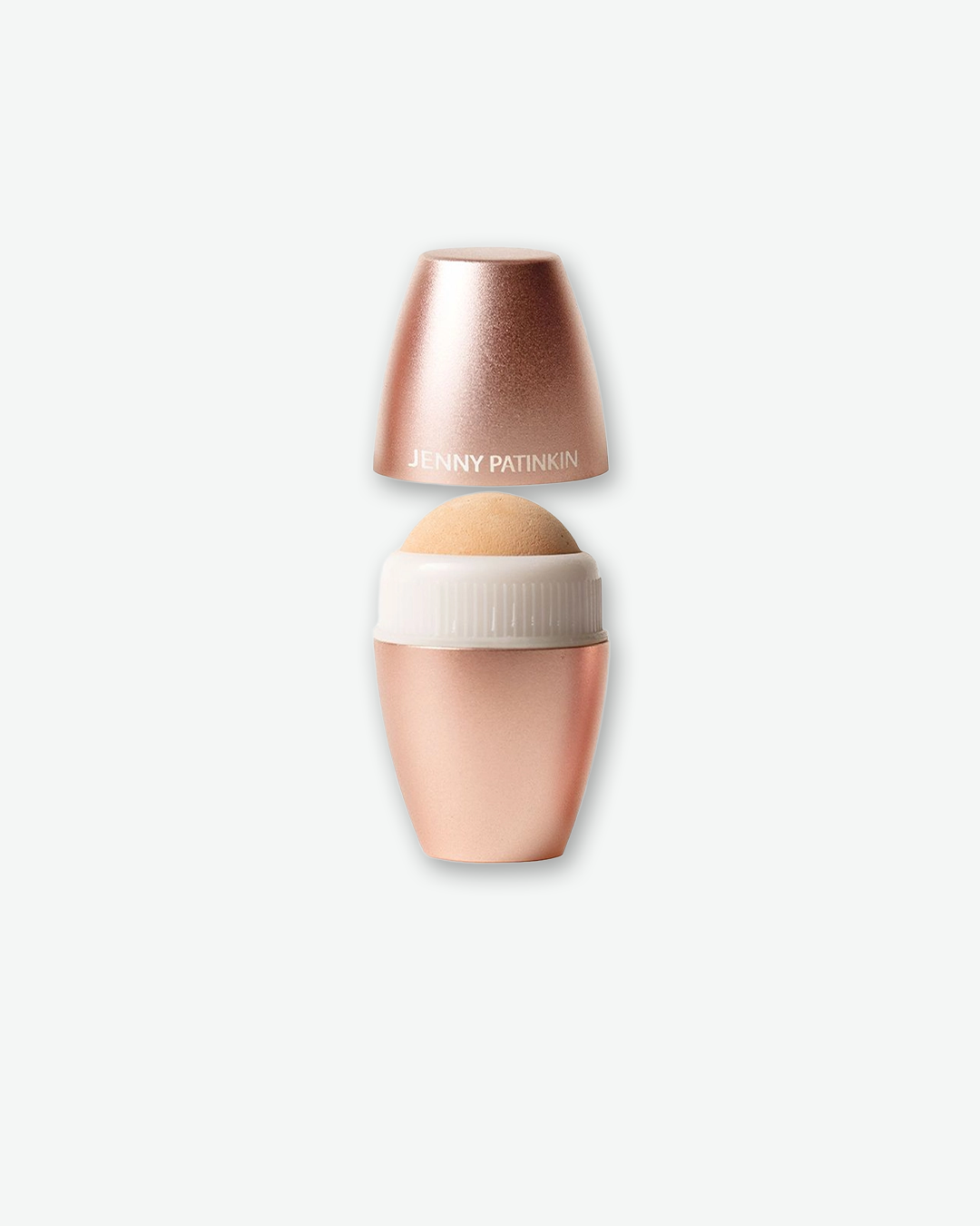 A product shot of an open Jenny Patinkin skin-toning tool centered against a plain light gray background. The metallic rose-gold cap, which features the brand name in white lettering, is floating slightly above the lower matching rose-gold barrel to reveal a smooth, rounded tan volcanic stone or sponge rolling ball at the top.