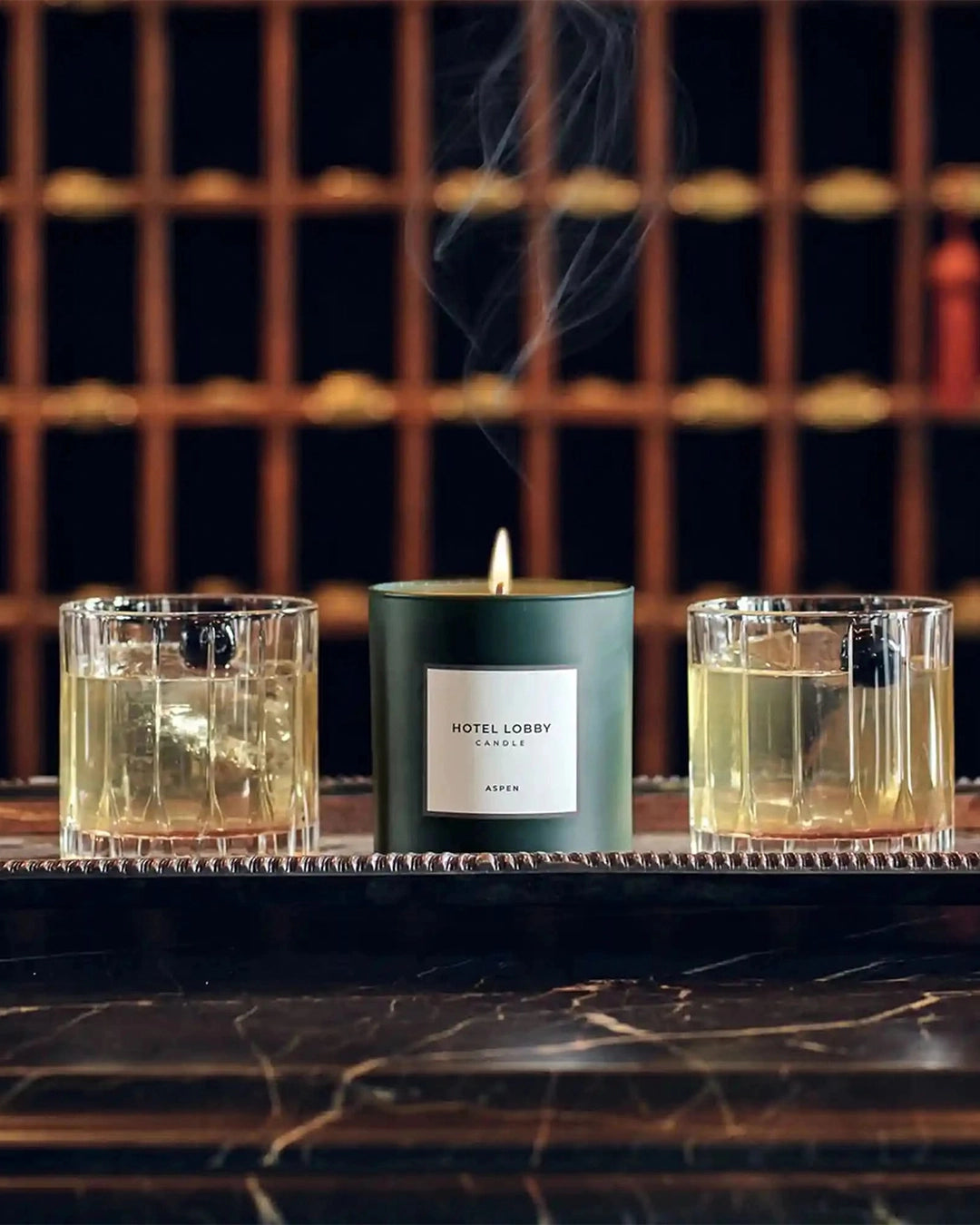 Two glasses of a yellowish liquid with a lit candle labeled 'Hotel Lobby' on a dark surface.