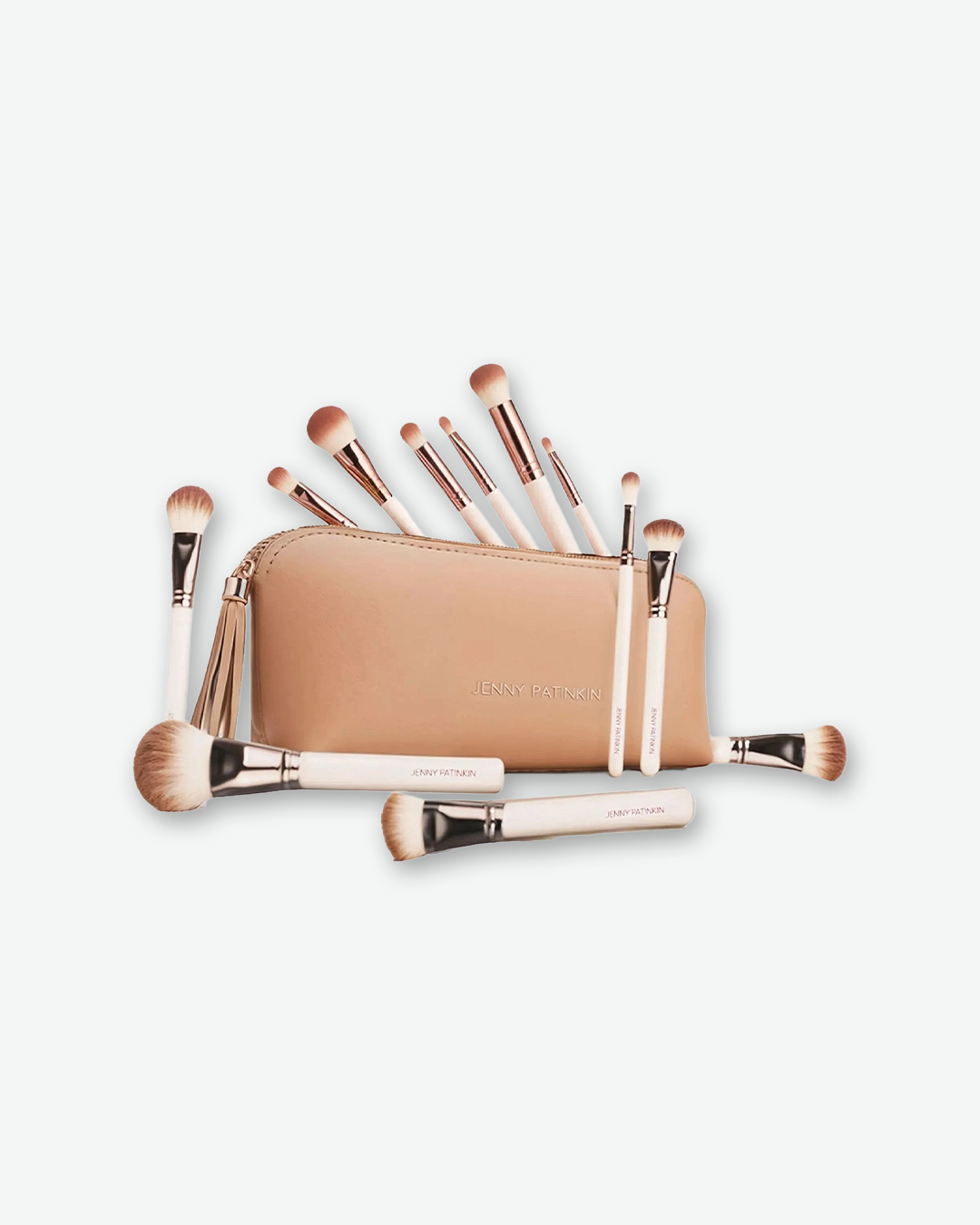 A product shot of a Jenny Patinkin professional makeup brush collection arranged around a tan cosmetic pouch on a light gray background. Several face and eye brushes with cream handles and rose-gold ferrules stand upright inside the opened pouch, while three larger complexion brushes rest horizontally on the surface in the foreground.