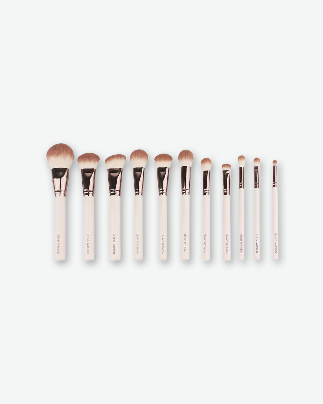 A product shot displaying a row of eleven Jenny Patinkin makeup brushes, arranged horizontally from largest to smallest against a plain light gray background. Each brush features a clean, cream-colored handle with "JENNY PATINKIN" printed vertically in small gold lettering, topped with a shiny rose-gold ferrule and dual-tone brown-and-white bristles.