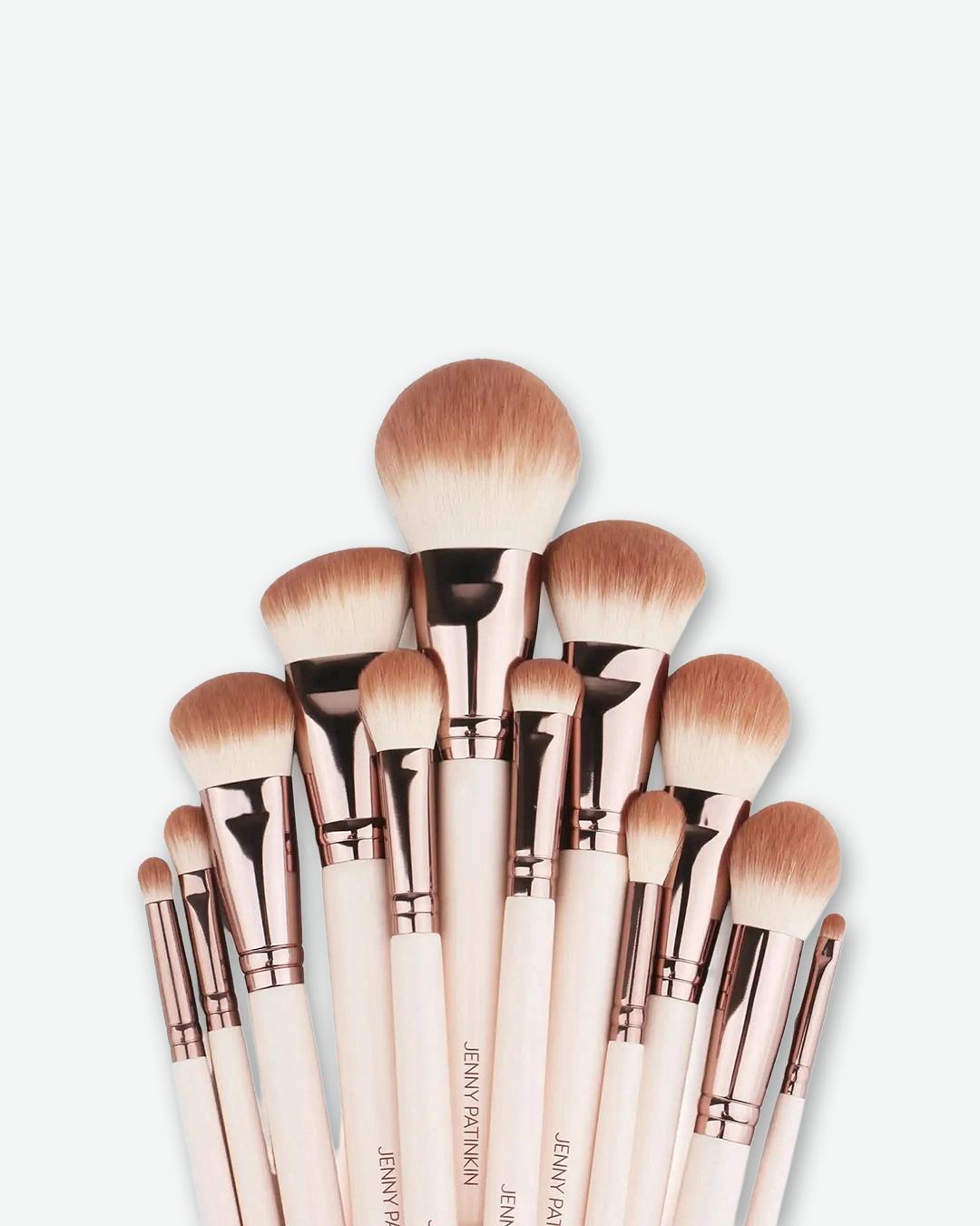A set of makeup brushes with pale pink handles, rose gold ferrules, and light brown bristles, arranged in a fan shape against a light gray background.