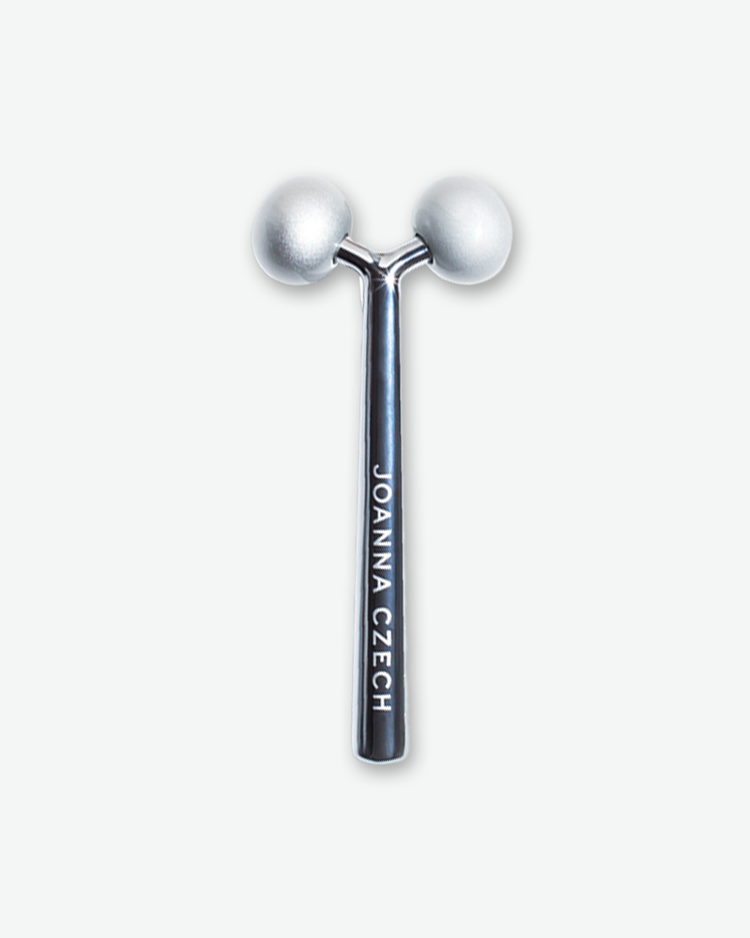 A product shot of a Joanna Czech Facial Massager, centered vertically against a plain light gray background. The tool features a sleek, reflective dark gray metallic handle with the name "JOANNA CZECH" printed vertically in clean white text. The top split-prong design holds two matte, textured silver rotating spheres designed to contour the face.
