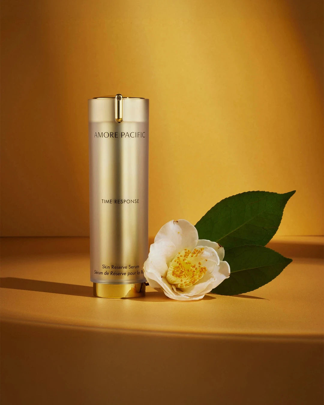 Gold cylindrical bottle of Amore Pacific Time Response Skin Reserve Serum bottle stood next to a white flower and green leaves on a warm yellow, shadowed background