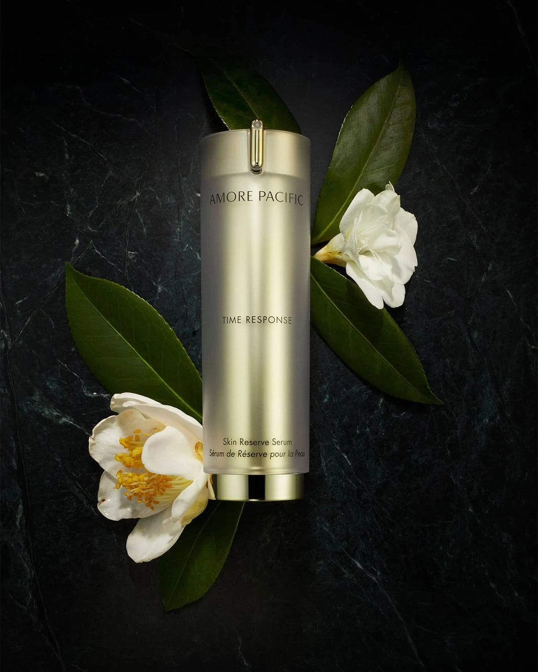 Gold Amore Pacific Time Response Skin Reserve Serum stood with white flowers and dark green leaves on a dark background