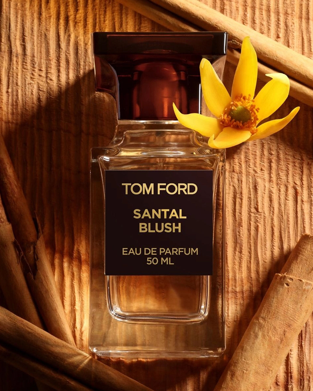 A warm macro shot of a Tom Ford Santal Blush 50ml perfume bottle lying flat against a background of deeply textured wood. It is framed closely by several whole cinnamon sticks and decorated with a vibrant, five-petaled yellow ylang-ylang flower resting on its upper right shoulder.