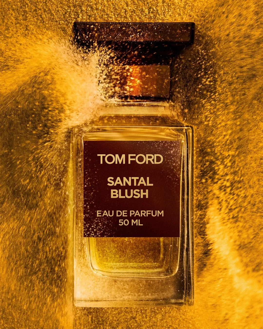 Artistic close-up campaign shot of Tom Ford Santal Blush Eau de Parfum. The architectural glass bottle is captured amidst a dynamic, swirling blast of fine golden-yellow powder that mimics a burst of fragrance spray, set against an intensely warm, backlit gold background.