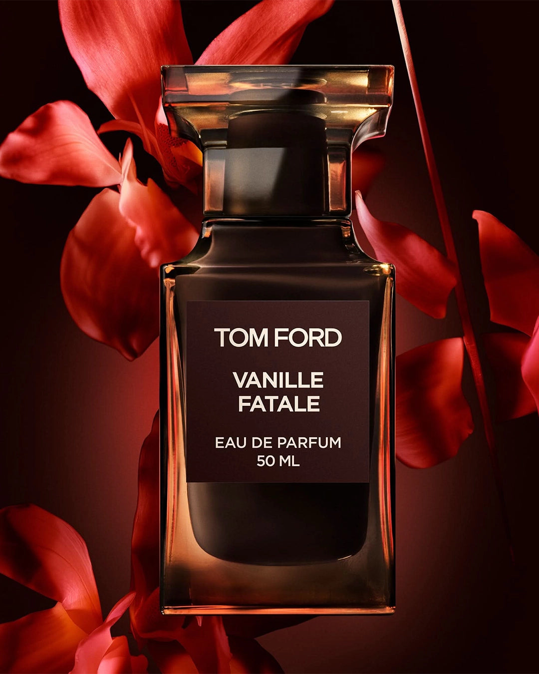 Close-up campaign shot of a Tom Ford Vanille Fatale perfume bottle. The dark glass bottle is backlit to glow with warm amber edges, positioned directly in front of large, velvety red flower petals that dramatic expand outward into a dark, shadowy red background.