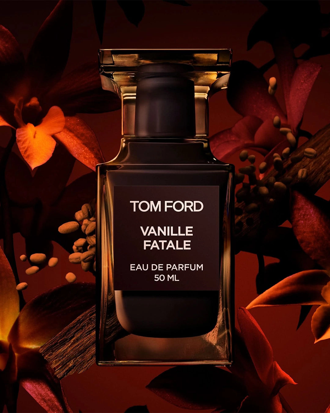 Dramatic holiday campaign imagery for Tom Ford Vanille Fatale Eau de Parfum. The dark amber glass bottle sits in the center, framed closely by shadowy, warm-toned deep red and orange orchid blossoms. Roasted coffee beans are scattered softly on the left side of the bottle, lit with moody, amber-toned lighting.